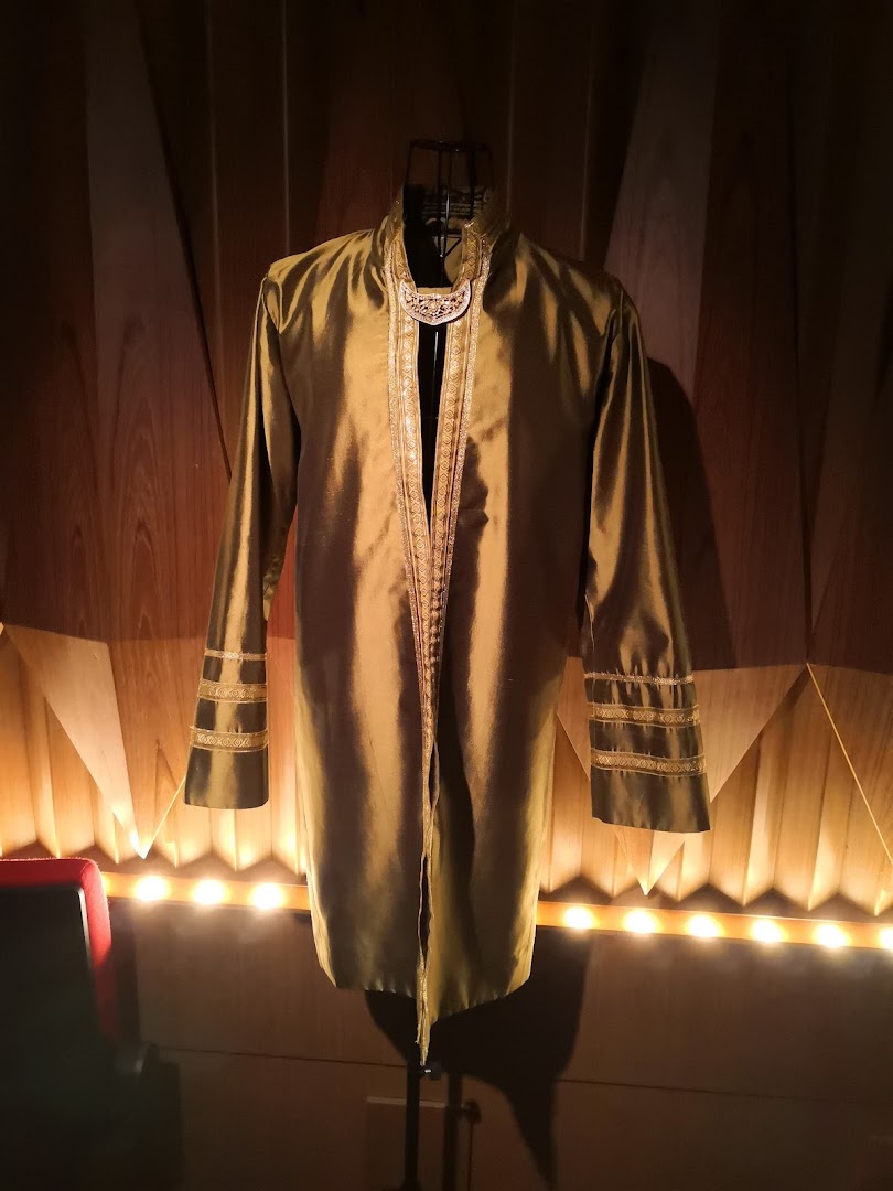 A golden-hued traditional garment with intricate trim displayed on a mannequin against a wooden wall.