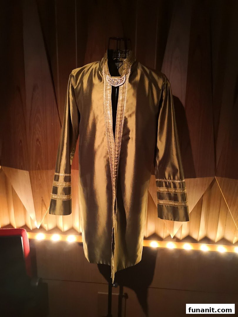 A golden-hued traditional garment with intricate trim displayed on a mannequin against a wooden wall.