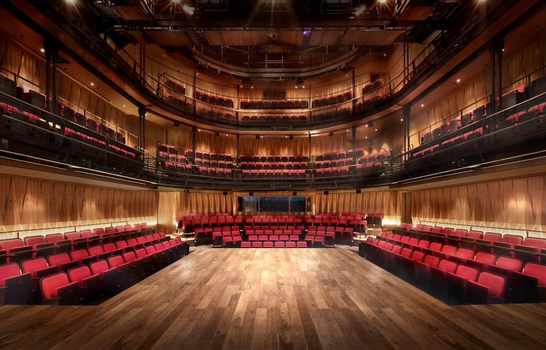 An empty theatre with multiple tiers of red seats, a wooden stage, and warm lighting.