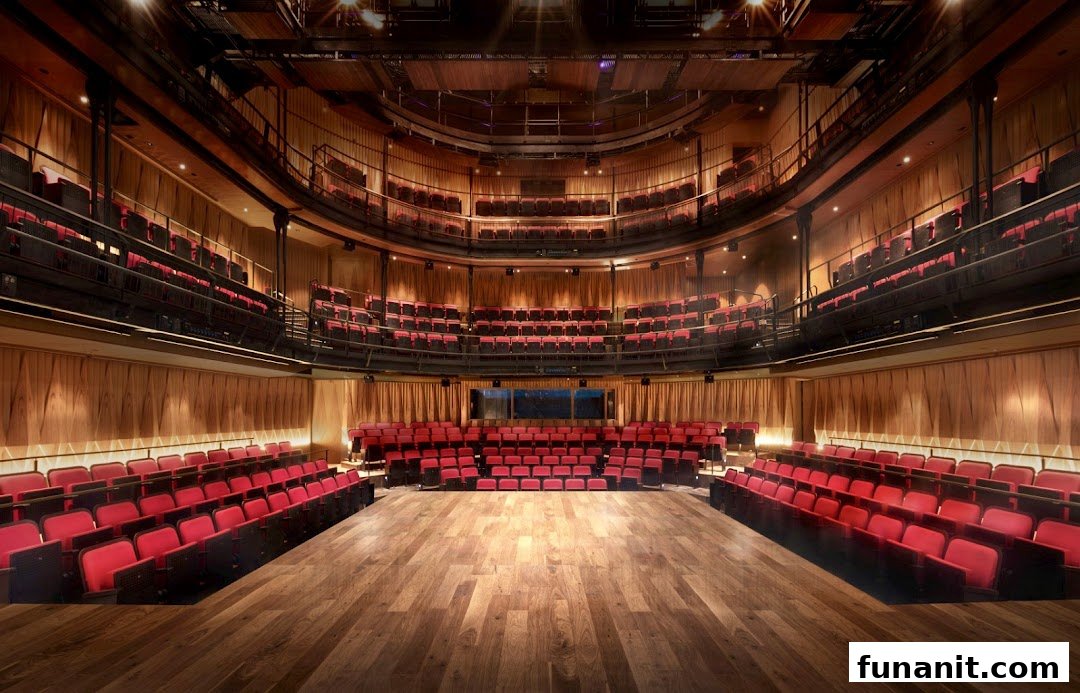 An empty theatre with multiple tiers of red seats, a wooden stage, and warm lighting.
