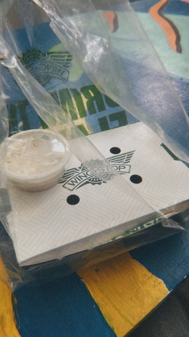 Wingstop packaging with a sauce cup, wrapped in plastic, on a patterned surface.