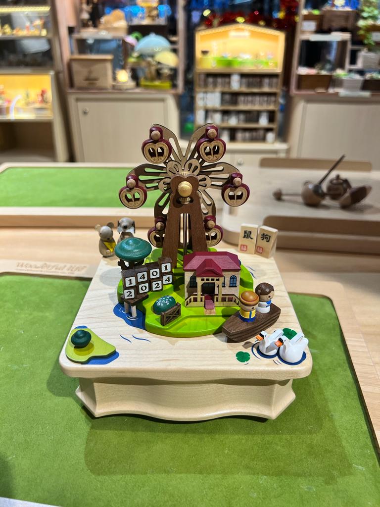 A detailed wooden music box featuring a Ferris wheel, house, and figures, displayed in a retail setting.