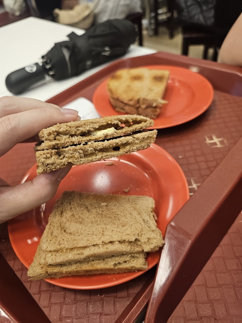 A hand holds a piece of kaya toast, revealing its sweet kaya and butter filling, with more toast on a red plate.