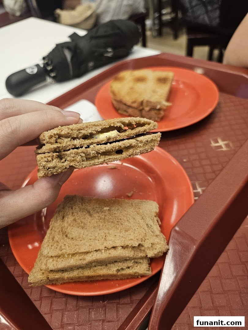 A hand holds a piece of kaya toast, revealing its sweet kaya and butter filling, with more toast on a red plate.