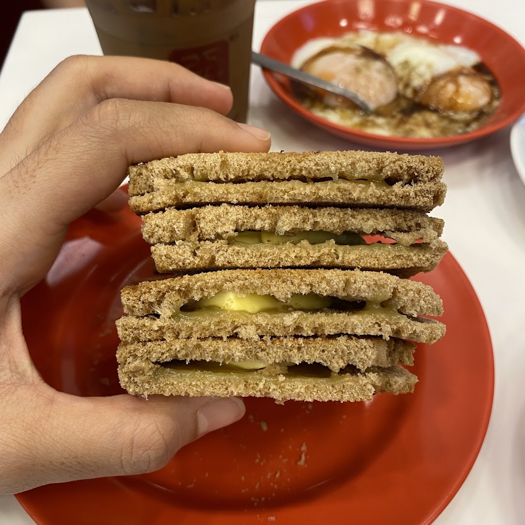 A hand holds a stack of kaya toast, with soft-boiled eggs and coffee visible in the background.