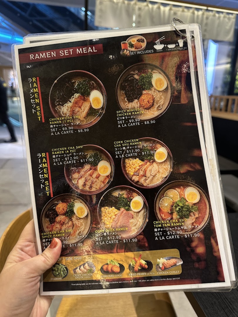 A hand holding a laminated menu displaying various ramen set meals and side dishes.