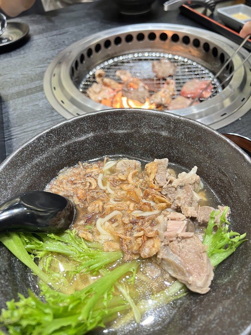 A close-up of a savory noodle bowl with meat and greens, with yakiniku grilling in the background.