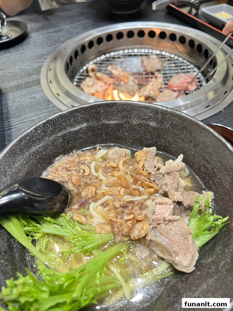 A close-up of a savory noodle bowl with meat and greens, with yakiniku grilling in the background.
