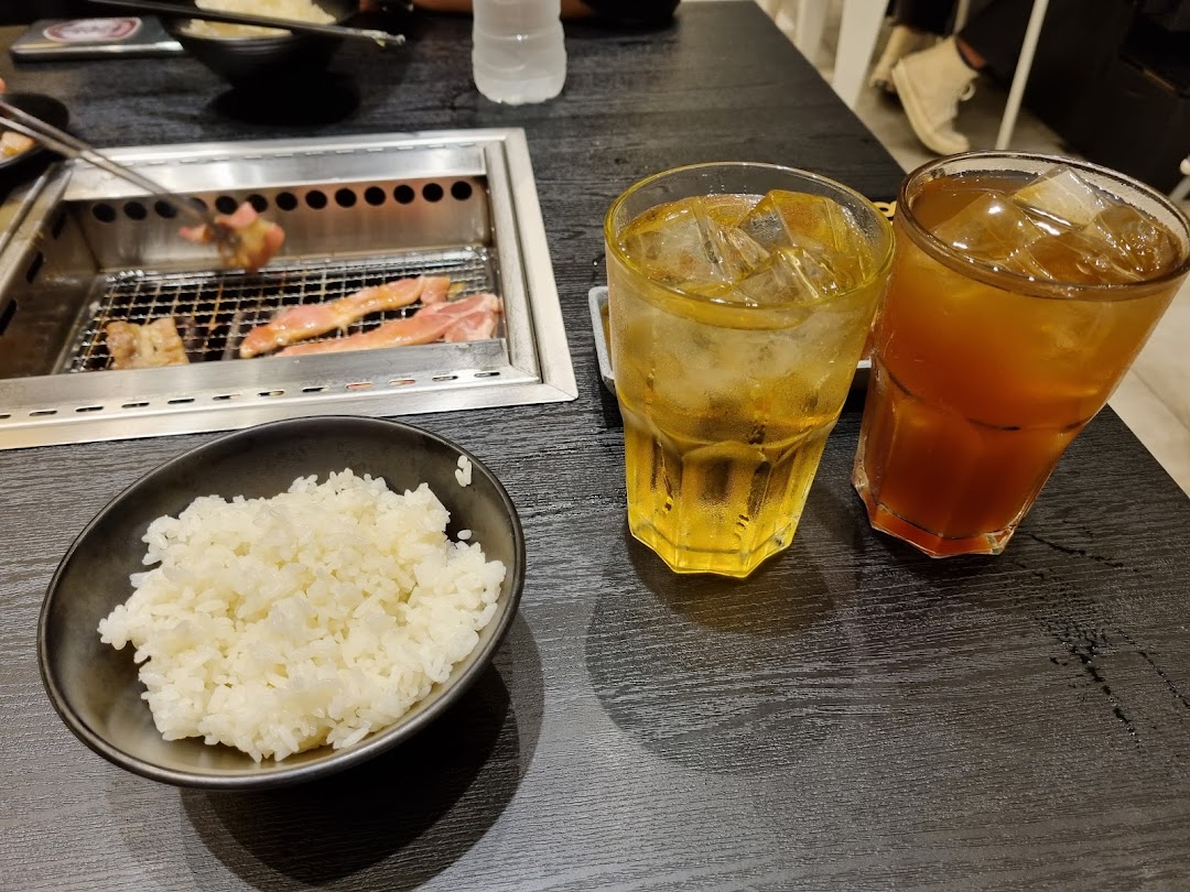 A close-up of a yakiniku grill with meat cooking, a bowl of rice, and two iced drinks on a black table.