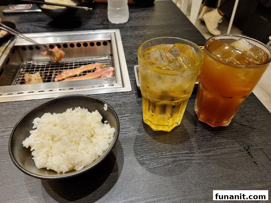 A close-up of a yakiniku grill with meat cooking, a bowl of rice, and two iced drinks on a black table.