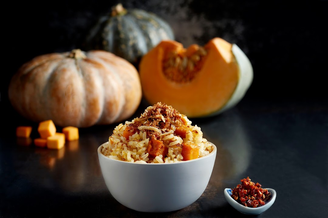 A steaming bowl of pumpkin rice topped with crispy anchovies and shallots, with fresh pumpkins in the background.