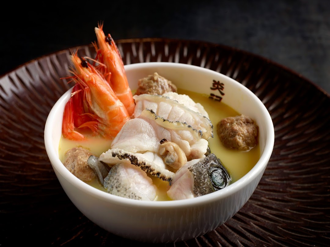 A close-up of a rich seafood soup with prawns, fish, and meatballs in a white bowl.