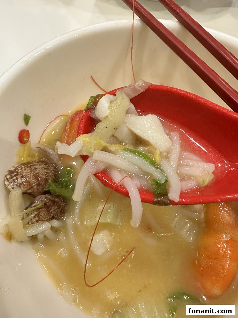 A close-up of a spoonful of gourmet seafood soup with noodles, vegetables, and seafood.