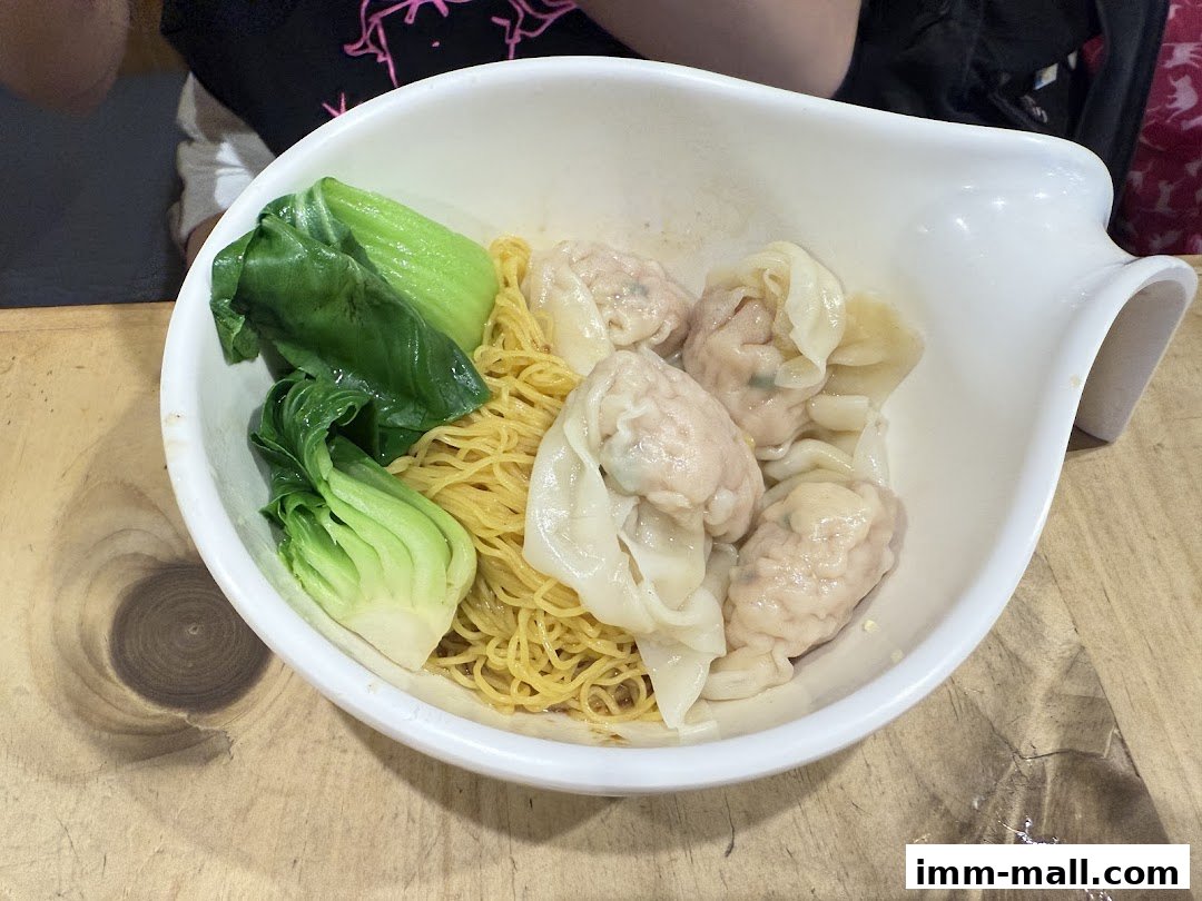 A bowl of wonton noodles with bok choy served in a white bowl on a wooden surface.
