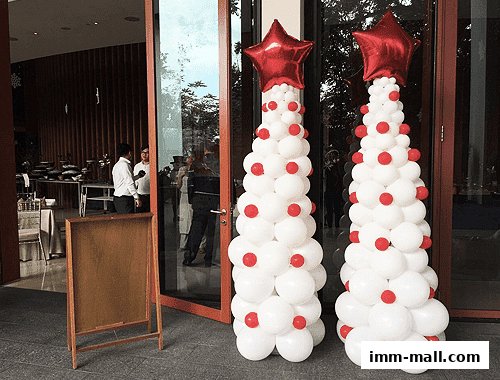 Two festive balloon trees with red stars stand outside a building entrance with people inside.