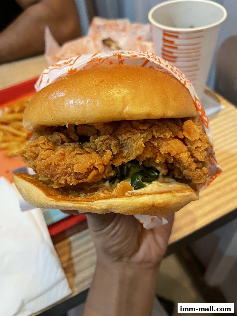 A hand holds a Popeyes chicken sandwich with fries and a drink in the background.
