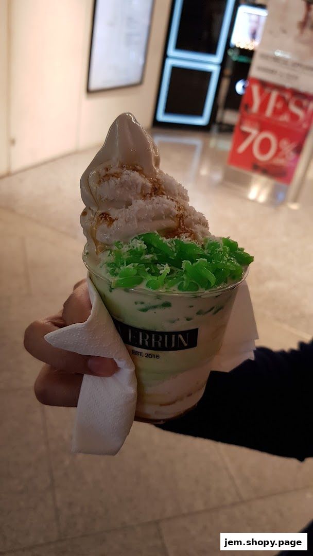 A hand holds a cup of dessert with ice cream, coconut, and green jelly.