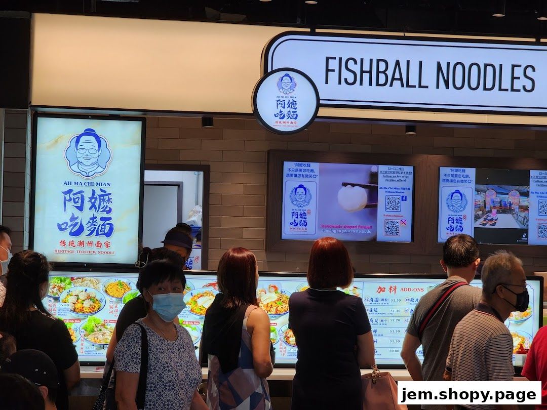 People queuing at a fishball noodle shop with digital menus displaying food and offers.