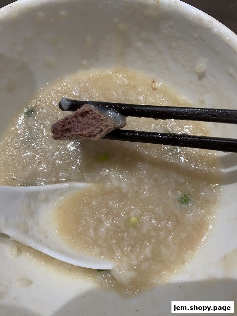 A close-up of a bowl of porridge with a piece of meat being held by chopsticks.