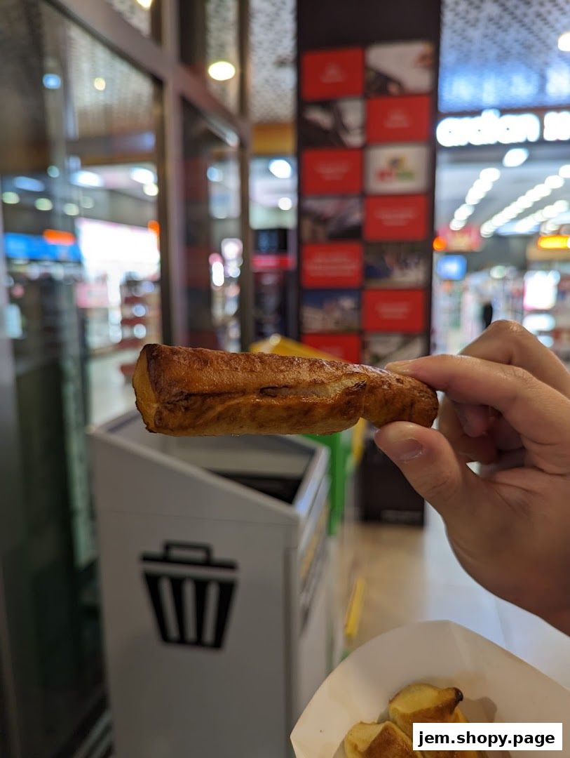A hand holds a pretzel stick in front of an Auntie Anne's pretzel shop.