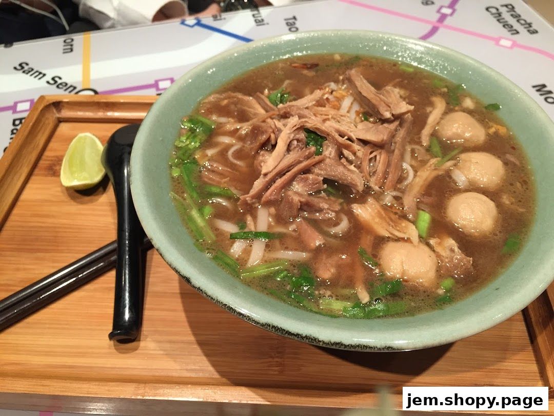 A bowl of duck noodle soup with meatballs, lime, and chopsticks on a wooden tray.