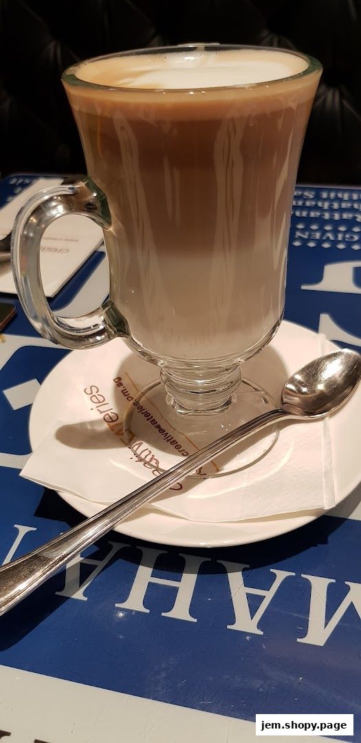 A tall glass of layered coffee with foam sits on a saucer with a spoon.