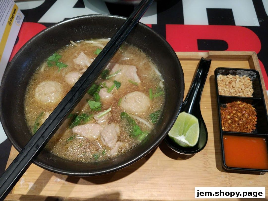 A steaming bowl of Thai noodle soup with meatballs, herbs, and a side of chili flakes, peanuts, and lime.