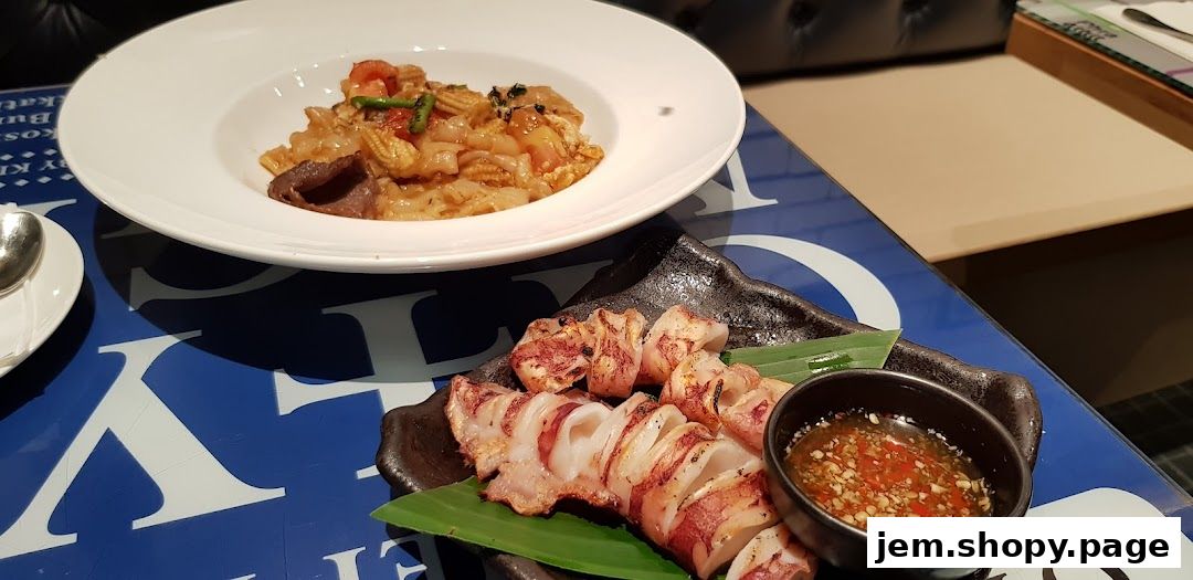 A plate of grilled squid with dipping sauce and a bowl of noodles on a blue patterned table.