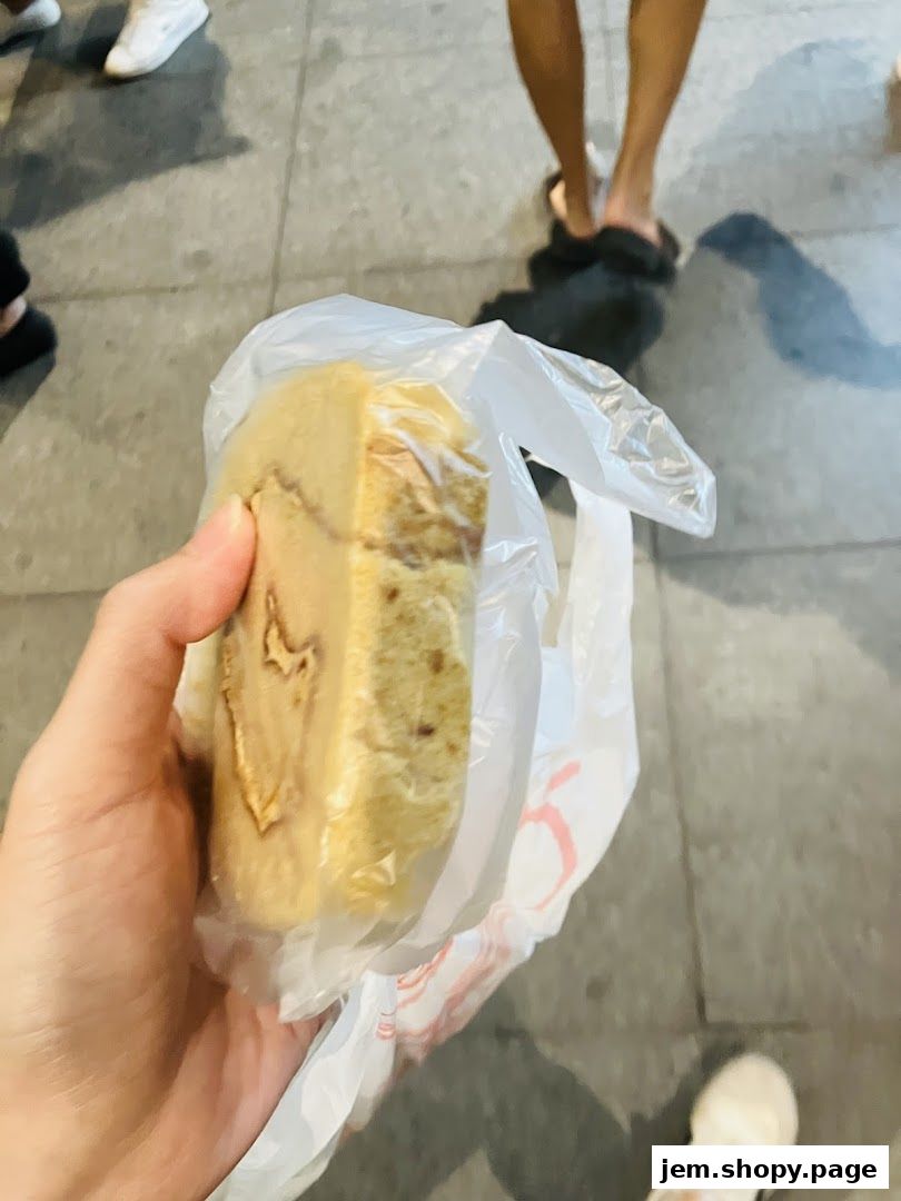 A hand holds a slice of cake in plastic packaging, with people walking in the background.
