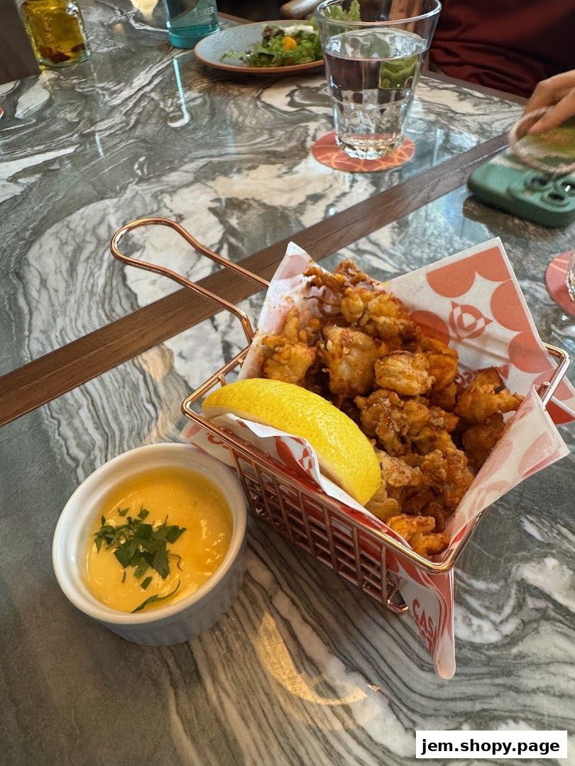 A basket of fried calamari with a lemon wedge and dipping sauce on a marble table.