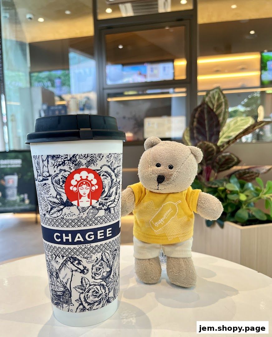 A CHAGEE branded cup and a teddy bear with a yellow shirt on a table.