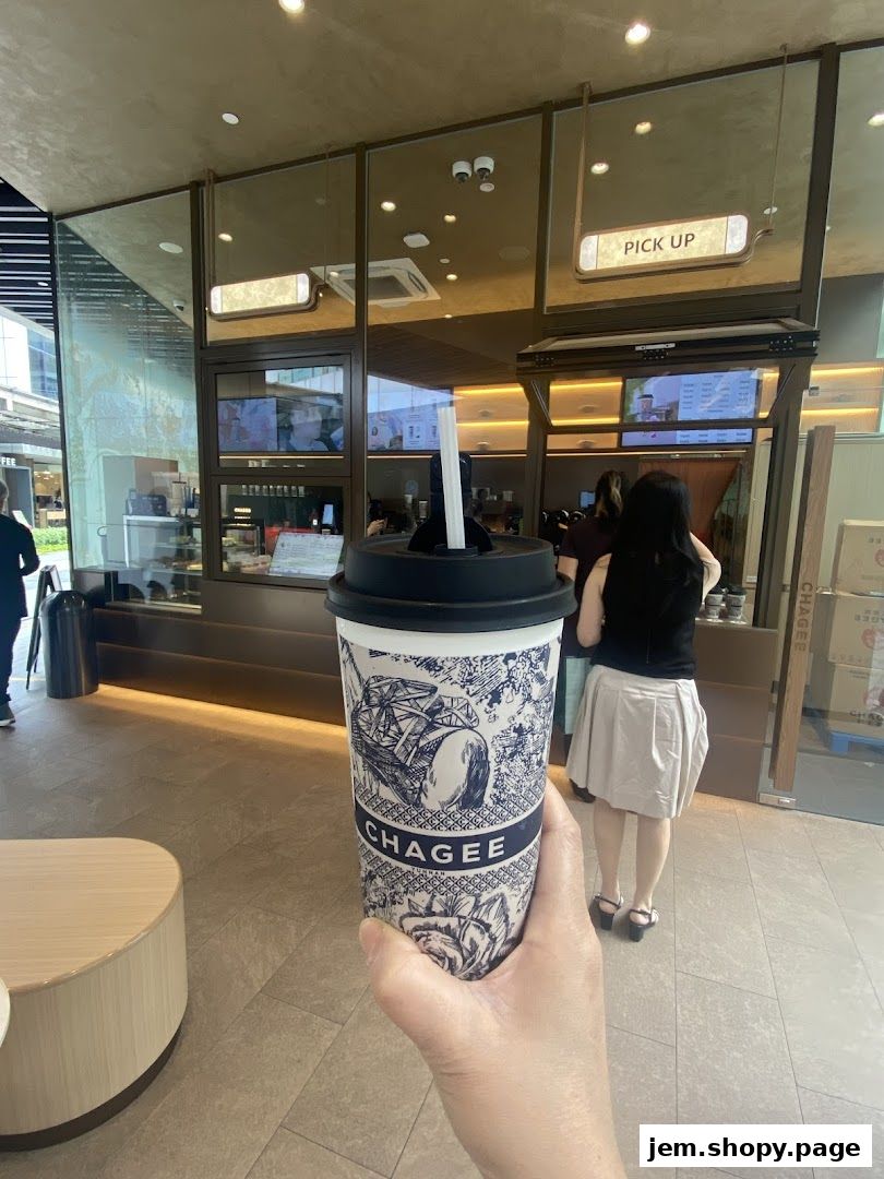 A hand holds a CHAGEE branded cup in front of a shop counter with a pick-up sign.