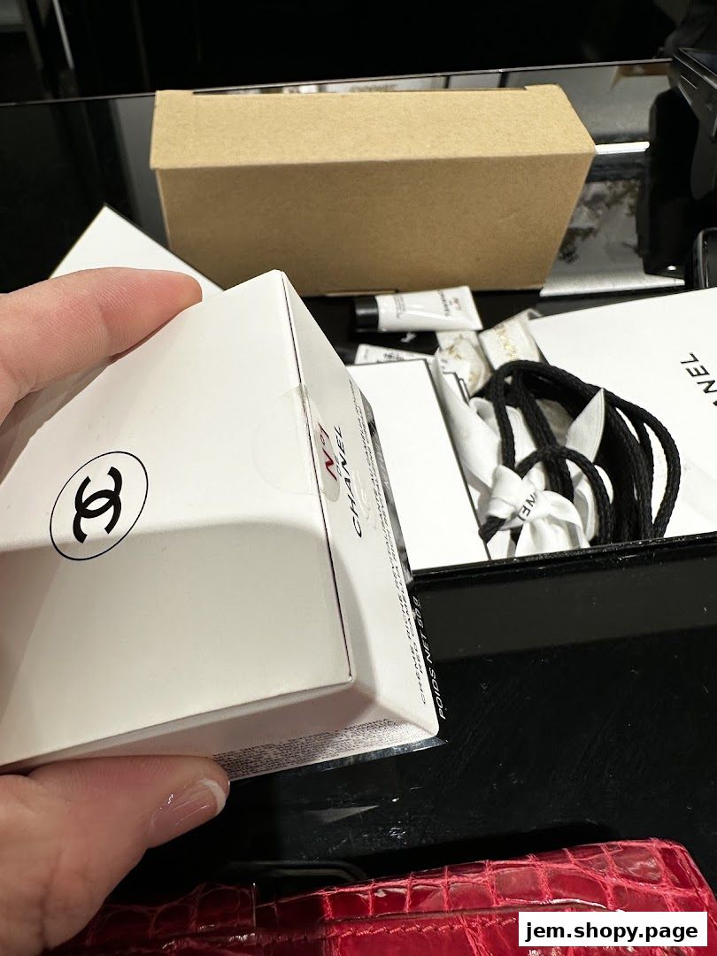 A hand holds a Chanel branded box with other Chanel products in the background.