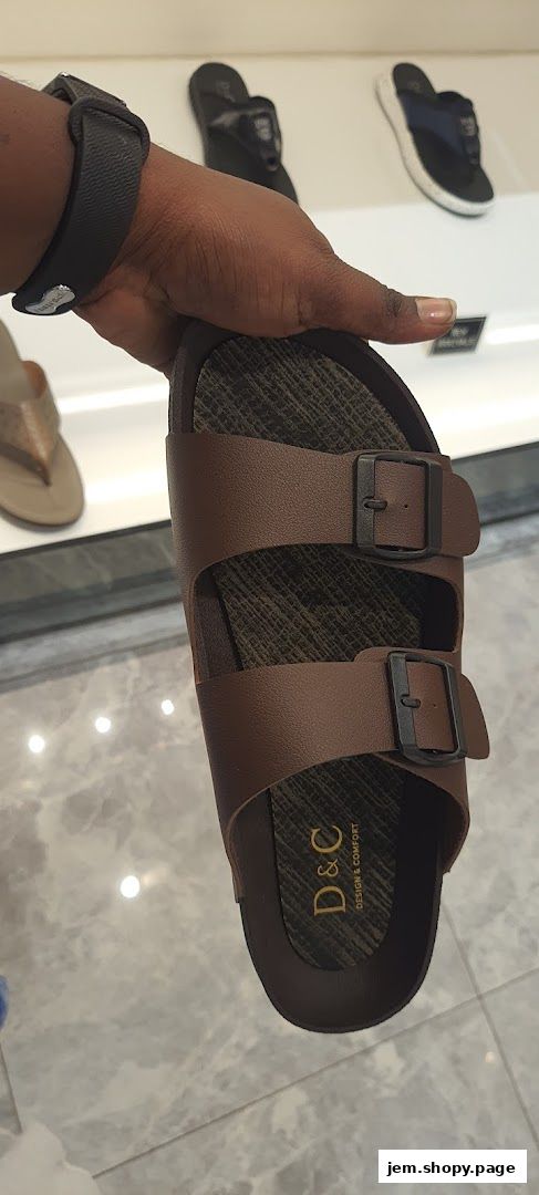 A hand holds a brown sandal with two buckles, showcasing the brand 'D&C'.