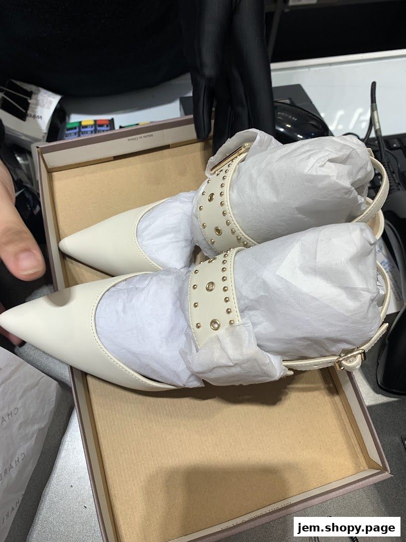 A pair of white pointed-toe heels with studded ankle straps in a box.