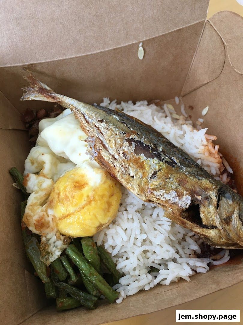 A close-up of a takeaway box filled with Nasi Lemak, featuring rice, fried fish, egg, and long beans.