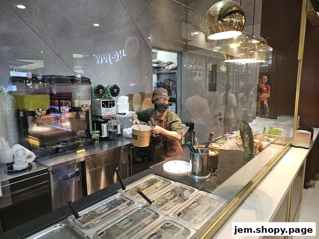 A food stall worker prepares a takeaway order behind a counter with drinks and food containers.