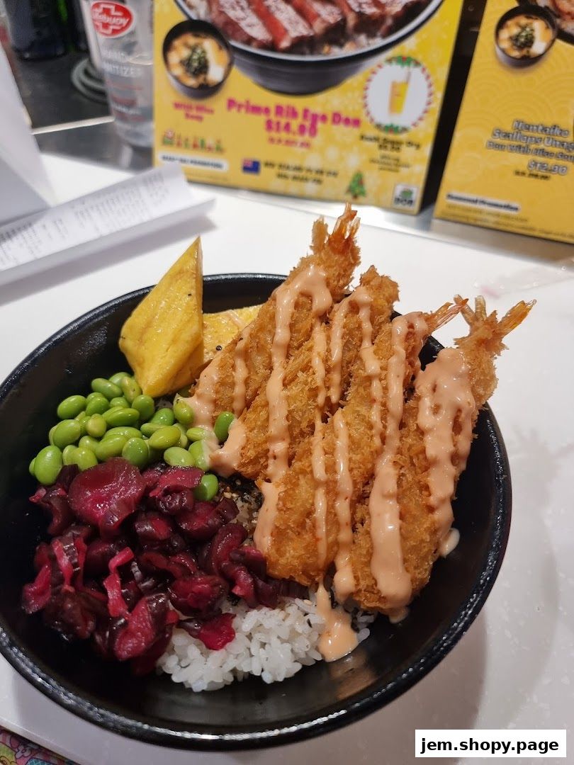 A close-up of a donburi bowl with fried shrimp, edamame, pickled vegetables, and rice.