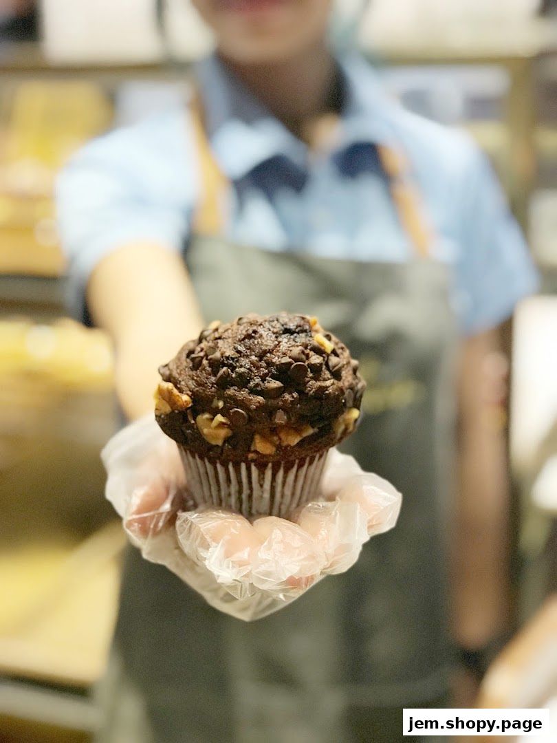A person in an apron holds out a chocolate chip and walnut muffin.