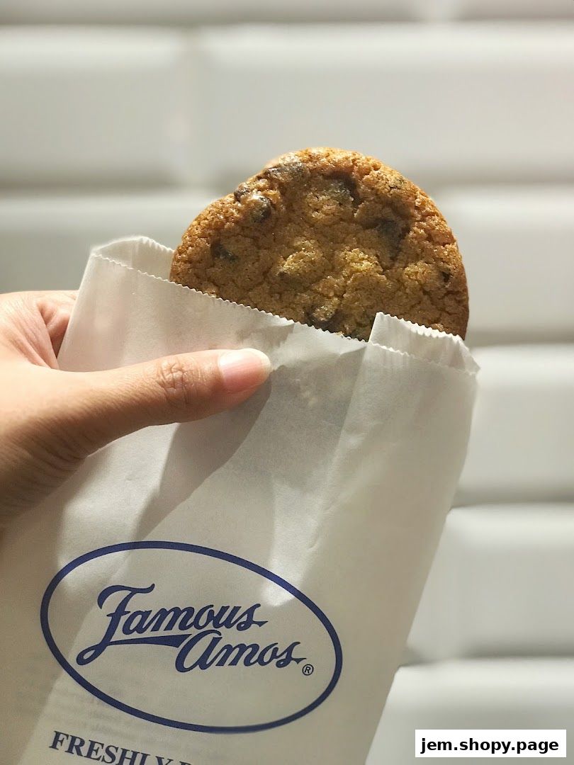 A hand holds a Famous Amos cookie in a branded paper bag.