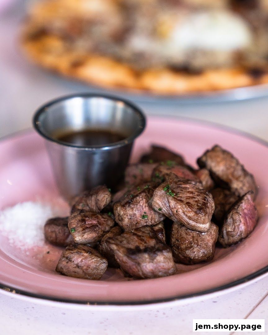 A plate of grilled steak bites with dipping sauce and salt, with a pizza in the background.