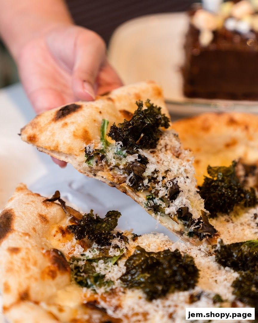 A hand holds a slice of pizza topped with kale and cheese, with a chocolate cake in the background.
