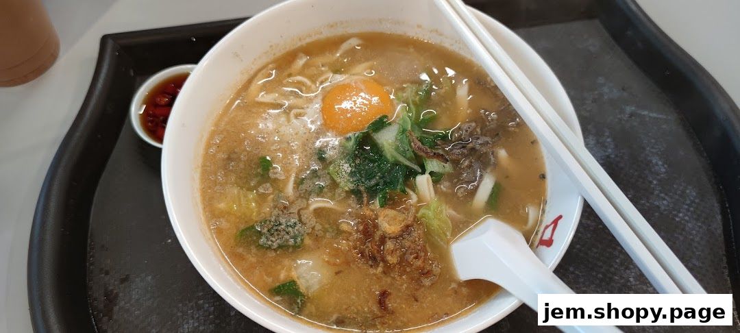 A bowl of Korean noodle soup with egg, vegetables, and meat, served with chopsticks and a side of chili sauce.