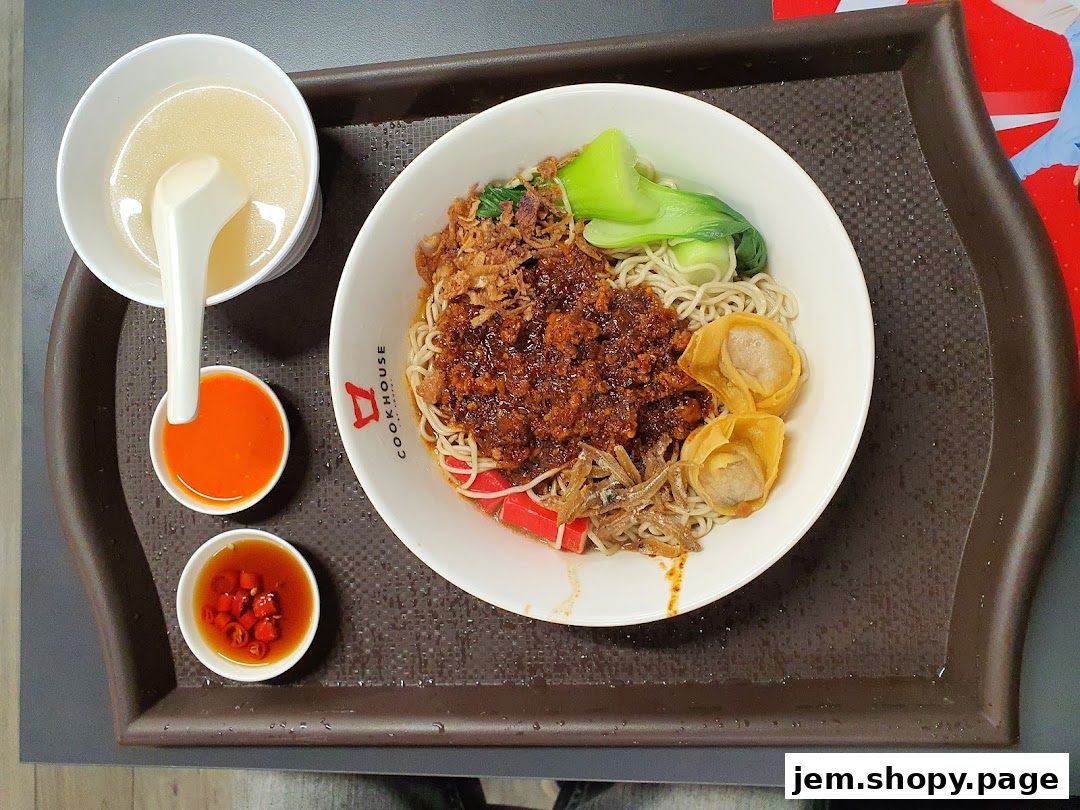 A bowl of noodles with minced meat, vegetables, and wontons, served with soup and chili sauce.