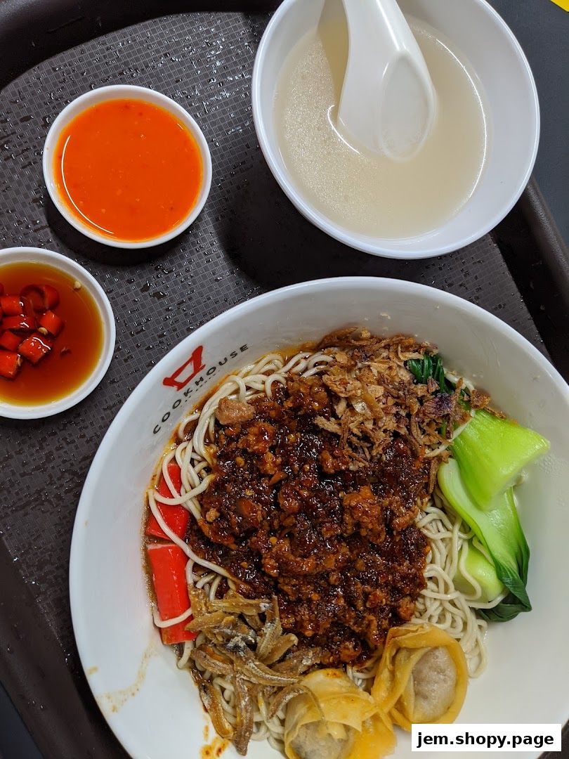 A bowl of noodles with minced meat, vegetables, and side dishes, served with soup and chili.