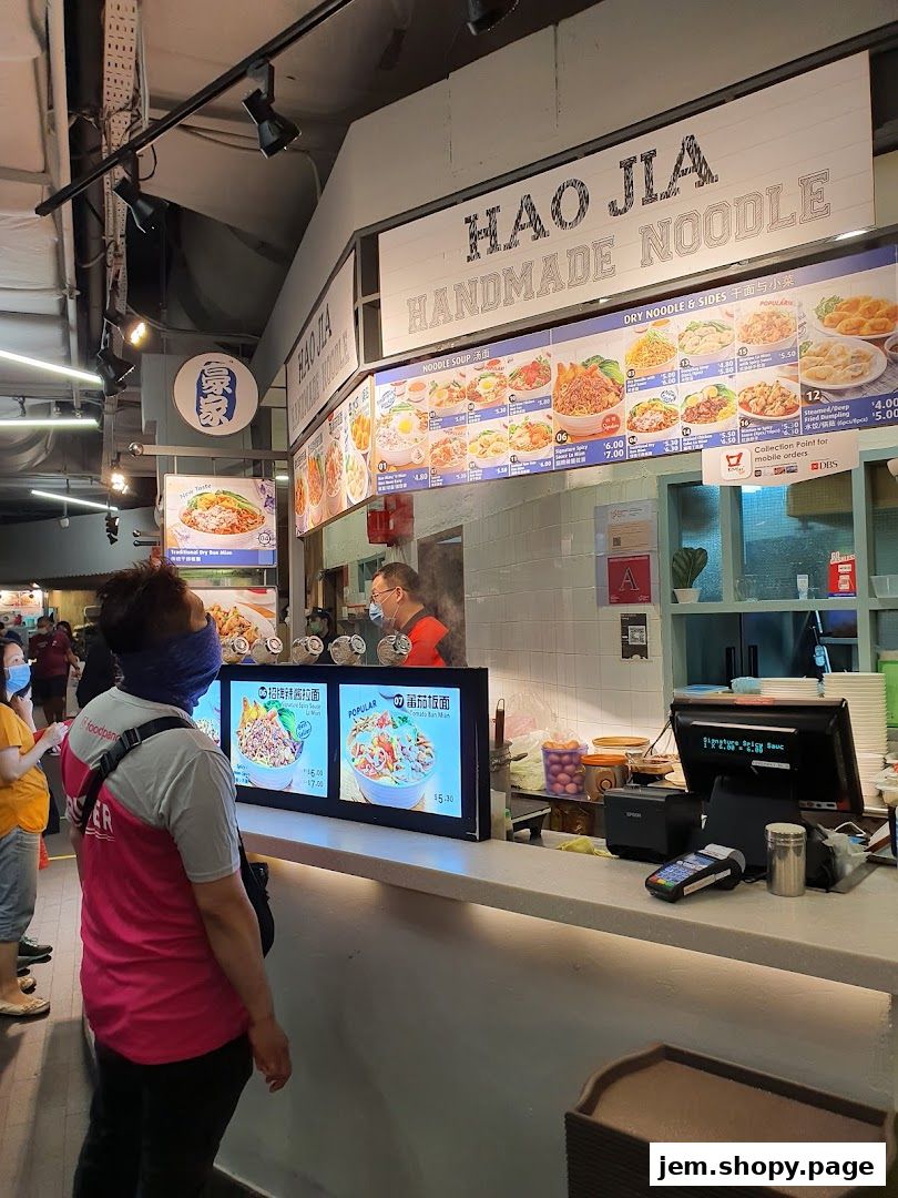 A food stall counter with a menu board displaying noodle dishes and a digital display.