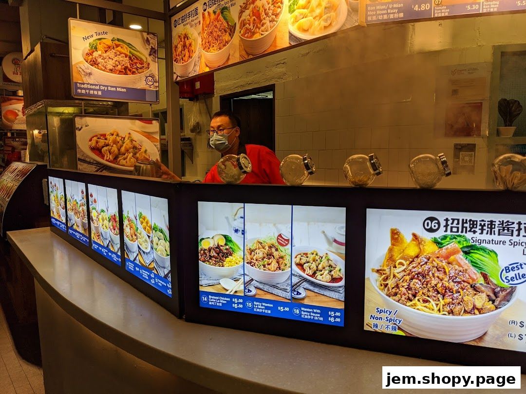 A food stall counter displaying various noodle dishes with prices and images.