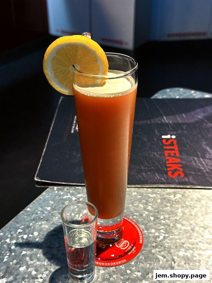 A tall glass of amber-colored drink with a lemon slice and a shot glass.