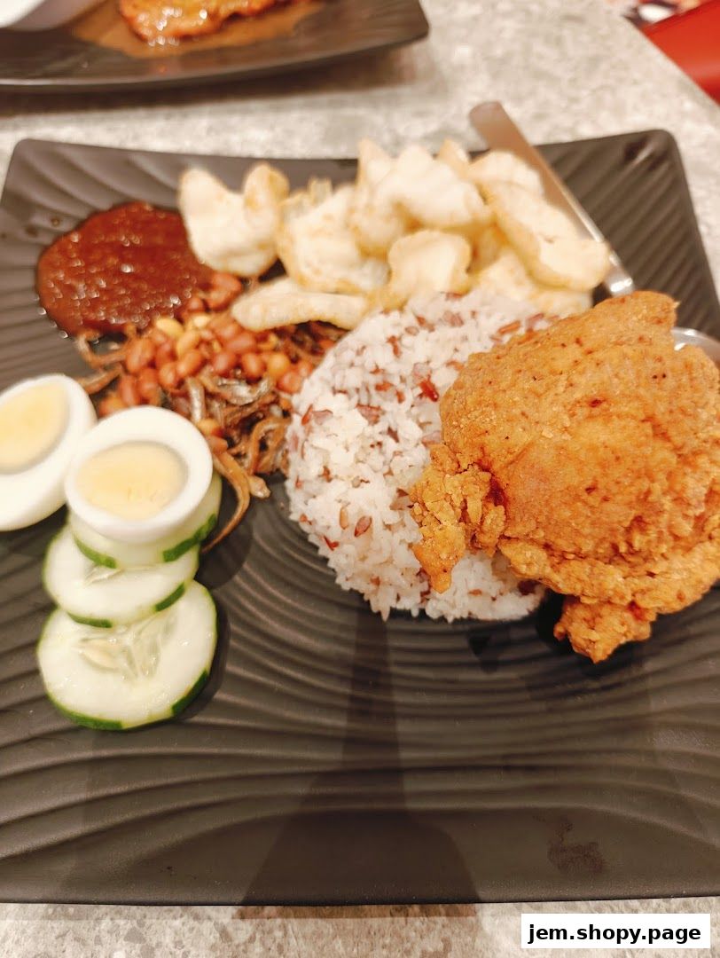 A plate of Nasi Lemak with fried chicken, rice, egg, cucumber, peanuts, and sambal.