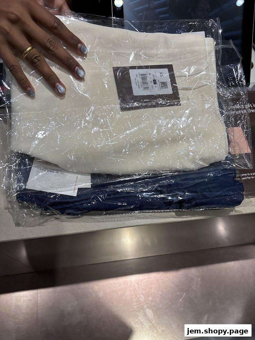 A hand with a ring and glittery nails points to a packaged beige top and blue pants.